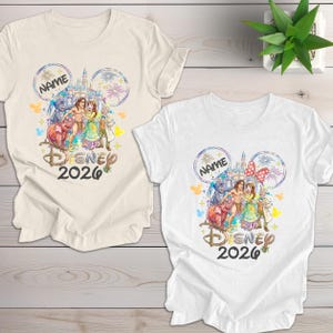 May include: Two t-shirts, one cream and one white, featuring a Disney-themed design. The design includes Disney characters, a castle, fireworks, and the text "DISNEY 2026". The design is framed by Mickey Mouse ears and the word "NAME".