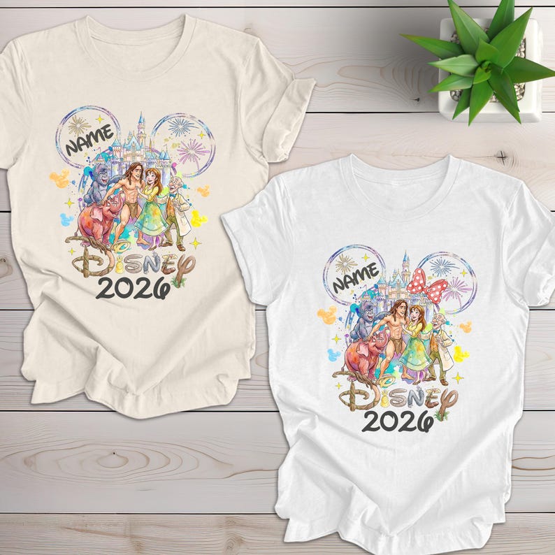 May include: Two t-shirts, one cream and one white, featuring a Disney-themed design. The design includes Disney characters, a castle, fireworks, and the text "DISNEY 2026". The design is framed by Mickey Mouse ears and the word "NAME".