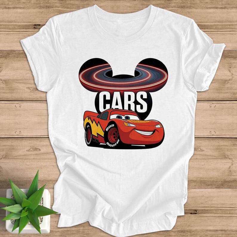 May include: White t-shirt with a graphic of Lightning McQueen from the film Cars. The design includes the word "CARS" and a stylized racetrack. The car is red with yellow and black accents.