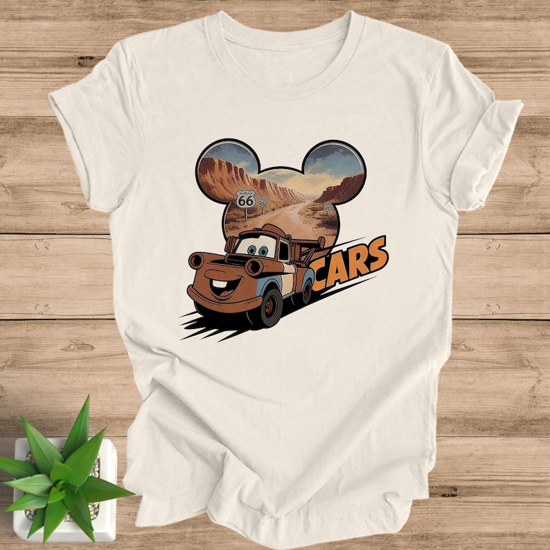 May include: Off-white t-shirt featuring a cartoon image of Mater from the movie Cars. The design includes a Route 66 sign and the word "Cars" in orange. The background is a desert landscape.