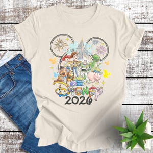 May include: Cream-colored t-shirt featuring a colorful graphic of Disney characters, including Woody, Buzz Lightyear, and others, with a castle and fireworks. The word "Disney" and the year "2020" are also displayed.