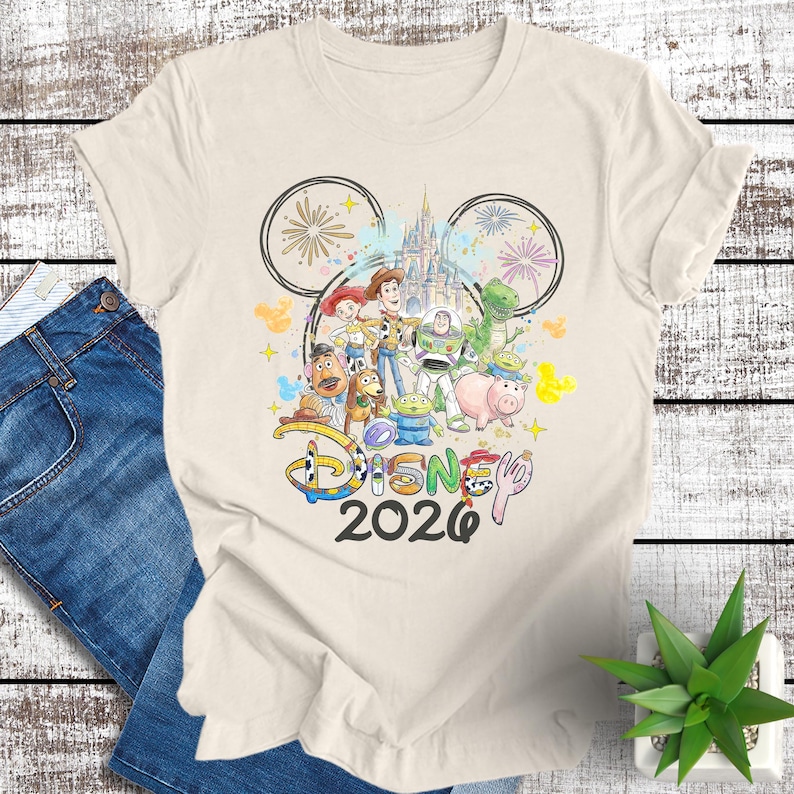 May include: Cream-colored t-shirt featuring a colorful graphic of Disney characters, including Woody, Buzz Lightyear, and others, with a castle and fireworks. The word "Disney" and the year "2020" are also displayed.