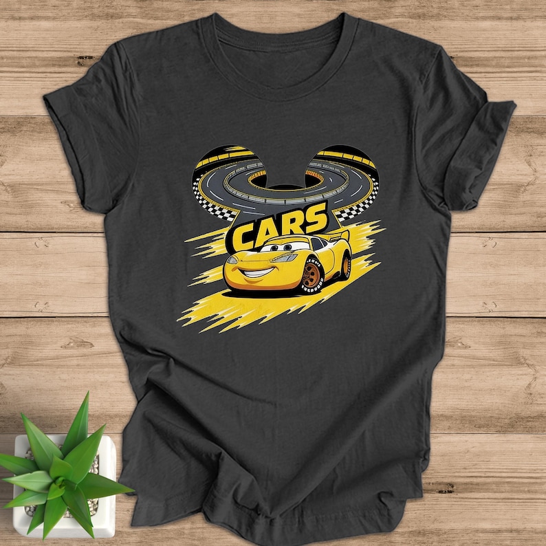May include: Dark grey t-shirt with a yellow cartoon race car and the word "CARS". The design includes a race track. The shirt is made of a soft material. The car has a smiling face. The design is in yellow and black.