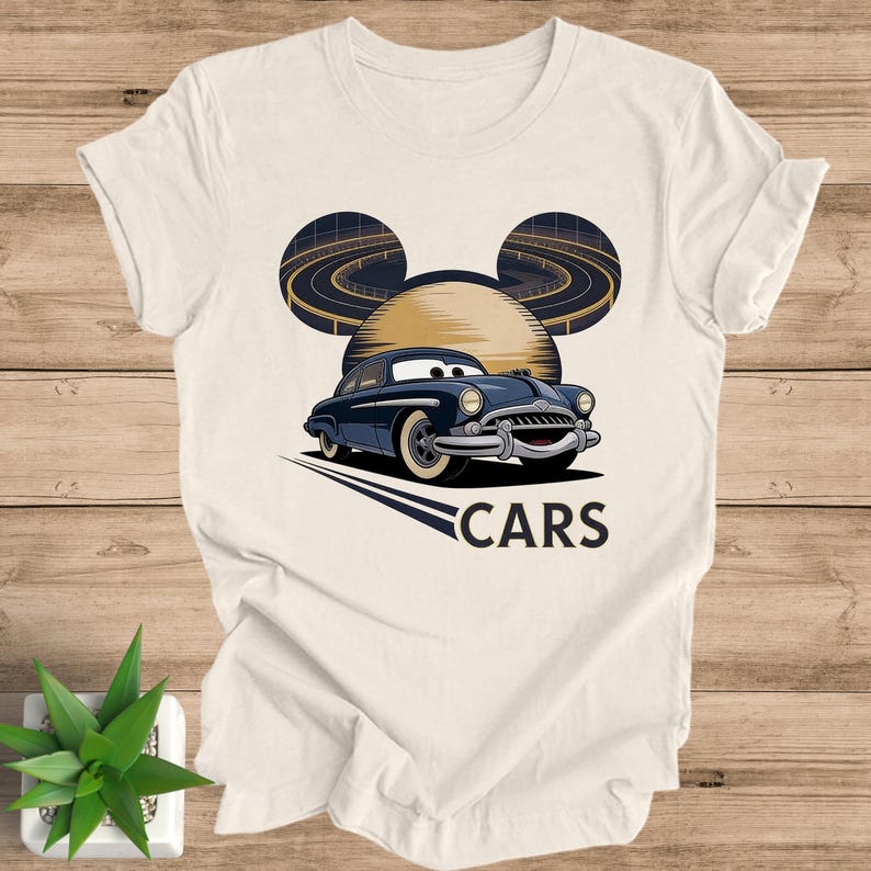 May include: Cream-colored t-shirt featuring a cartoon car design. The design includes a navy blue car with white tires, set against a backdrop of a stylized sun and race tracks, with the word "CARS" below.