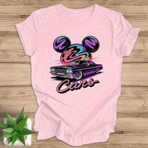 May include: A light pink t-shirt with a graphic of a purple car and the word "Cars". The design includes a stylized sun and Mickey Mouse ears. The t-shirt is made of a soft material.