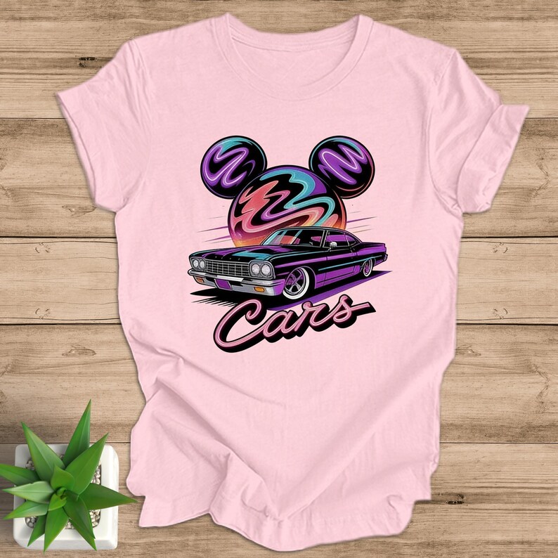 May include: A light pink t-shirt with a graphic of a purple car and the word "Cars". The design includes a stylized sun and Mickey Mouse ears. The t-shirt is made of a soft material.