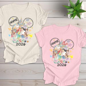May include: Two t-shirts, one cream and one pink, featuring a Disney-themed design. The design includes Disney characters, a castle, fireworks, and the text "NAME" and "DISNEY 2026" in a Mickey Mouse ear outline.