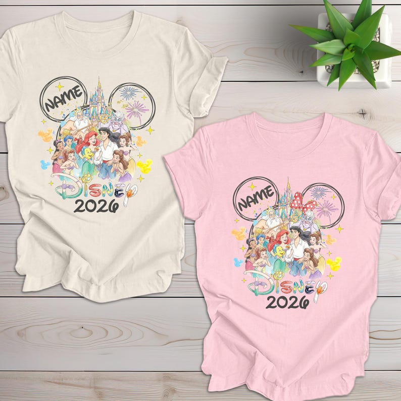 May include: Two t-shirts, one cream and one pink, featuring a Disney-themed design. The design includes Disney characters, a castle, fireworks, and the text "NAME" and "DISNEY 2026" in a Mickey Mouse ear outline.