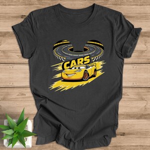 May include: Dark gray t-shirt featuring a yellow cartoon race car with the word "CARS" above it. The car is smiling and set against a yellow and black race track design. The shirt is made of a soft material.