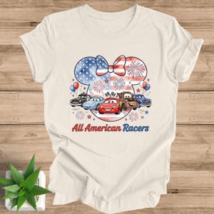 May include: Cream-colored t-shirt featuring a patriotic design with a bow and fireworks. The design includes cartoon cars and the text "All American Racers". The shirt is made of a soft material.