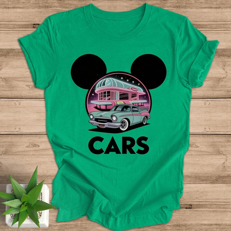May include: Green t-shirt featuring a retro car design with a diner and the word "CARS" in bold letters. The design includes a light blue car, a pink diner, and black Mickey Mouse ears.
