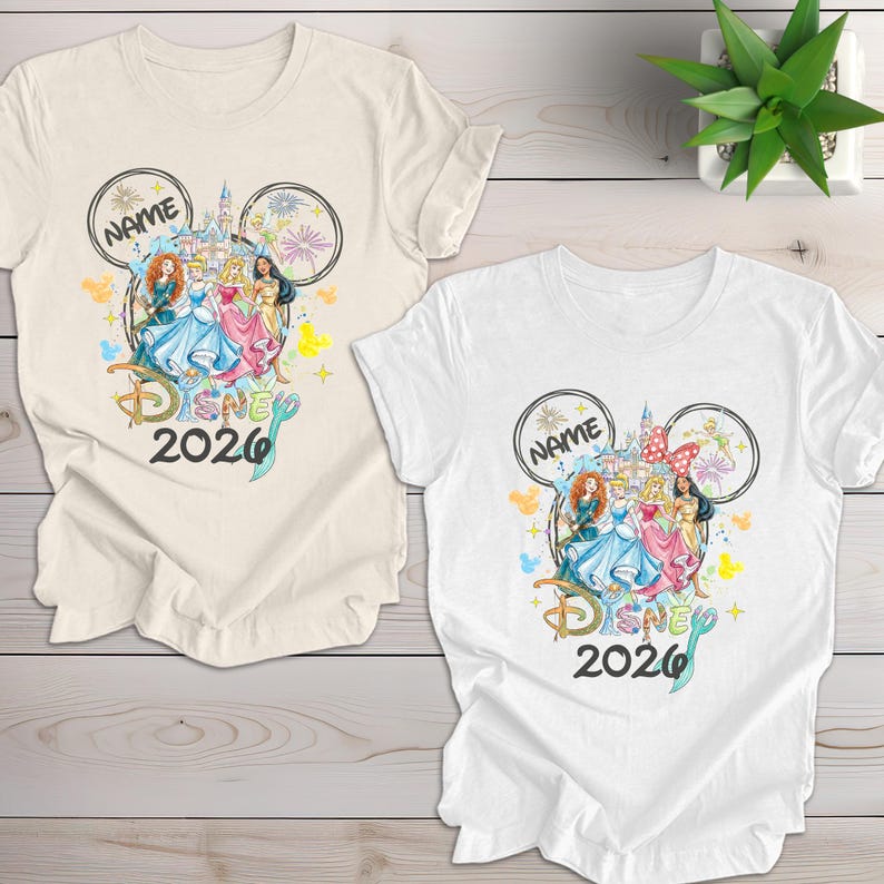 May include: Two t-shirts, one cream and one white, featuring a Disney-themed design. The design includes Disney princesses, a castle, fireworks, and the text "Disney 2026". A Mickey Mouse ear outline frames the design, with space for a name.