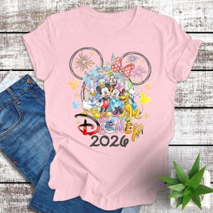 May include: A light pink t-shirt featuring a colorful Disney design. The design includes Mickey and Minnie Mouse, Donald and Daisy Duck, Goofy, and Pluto, with a castle and fireworks. The text "Disney 2026" is also included.
