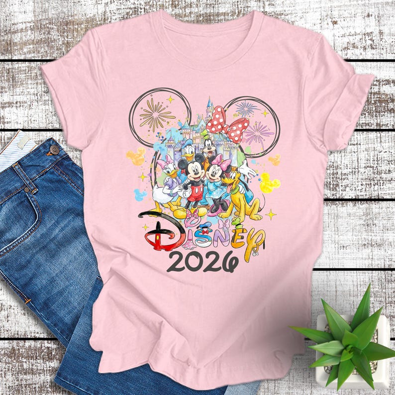 May include: A light pink t-shirt featuring a colorful Disney design. The design includes Mickey and Minnie Mouse, Donald and Daisy Duck, Goofy, and Pluto, with a castle and fireworks. The text "Disney 2026" is also included.