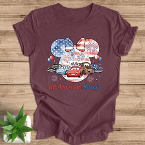 May include: Burgundy t-shirt featuring a patriotic design with a Minnie Mouse silhouette, a bow in red, white, and blue, and fireworks. Below the silhouette are cartoon race cars and the text "All American Racers."