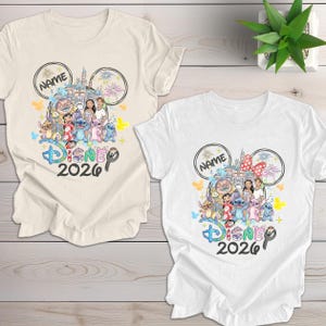 May include: Two t-shirts, one cream and one white, featuring a colorful Disney design. The design includes Disney characters, a castle, fireworks, and the text "Disney 2026". A name is written inside a Mickey Mouse ear.