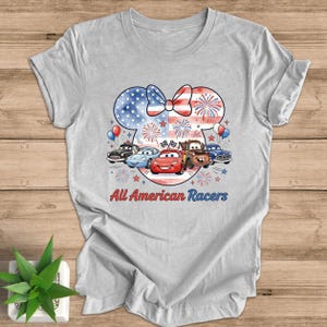 May include: A light gray t-shirt featuring a patriotic design with a Minnie Mouse silhouette, a bow, and fireworks. Below the design are cartoon cars and the text "All American Racers".