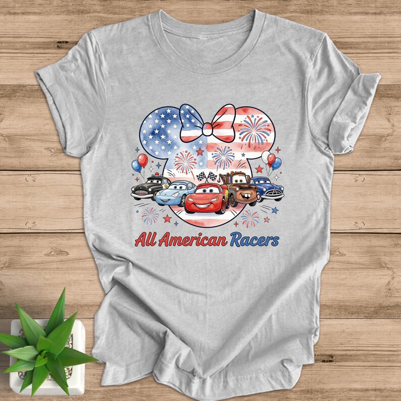 May include: A light gray t-shirt featuring a patriotic design with a Minnie Mouse silhouette, a bow, and fireworks. Below the design are cartoon cars and the text "All American Racers".