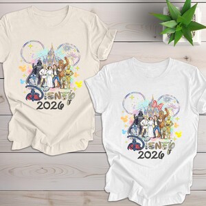 May include: Two t-shirts, one cream and one white, featuring a colorful graphic. The design includes Disney characters, a castle, fireworks, and the text "Disney 2026" within Mickey Mouse ears. The shirts are on a light wood surface.