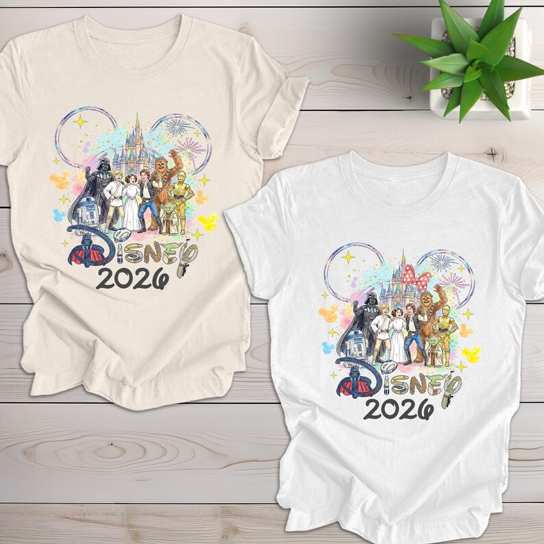 May include: Two t-shirts, one cream and one white, featuring a colorful graphic. The design includes Disney characters, a castle, fireworks, and the text "Disney 2026" within Mickey Mouse ears. The shirts are on a light wood surface.