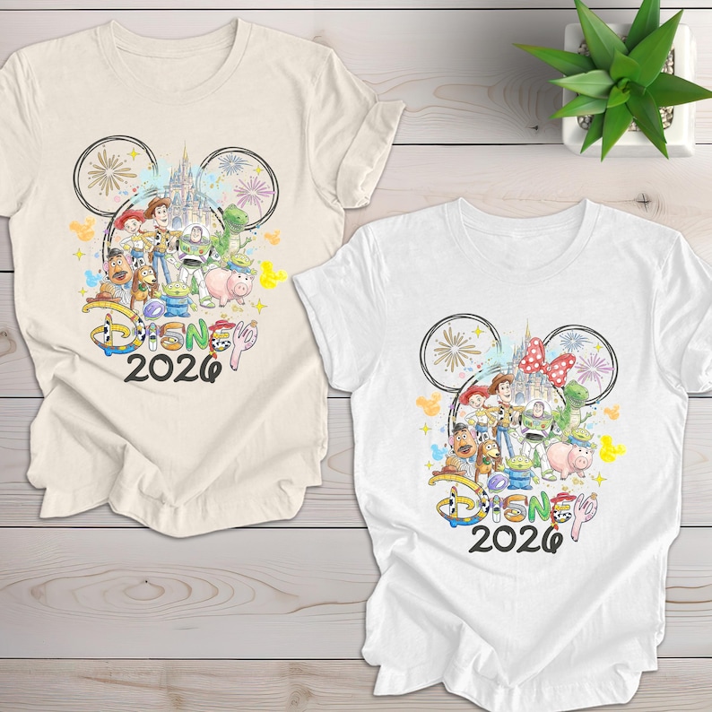 May include: Two white and cream-colored t-shirts featuring a colorful Disney graphic. The graphic includes Disney characters, a castle, and the text "Disney 2026" within a Mickey Mouse ear outline.