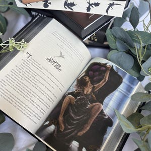May include: An open hardcover book titled Iron Flame by Rebecca Yarros, featuring a printed illustration of a person sitting on a dark ornate chair. The page displays Chapter Forty-Eight with a small dragon icon. Stacked books and eucalyptus leaves surround it.