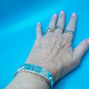 May include: A silver bracelet engraved with the word "Mom" and the phrase "I love my FAMILY". The bracelet is on a wrist with rings on the fingers. The background is a solid blue.
