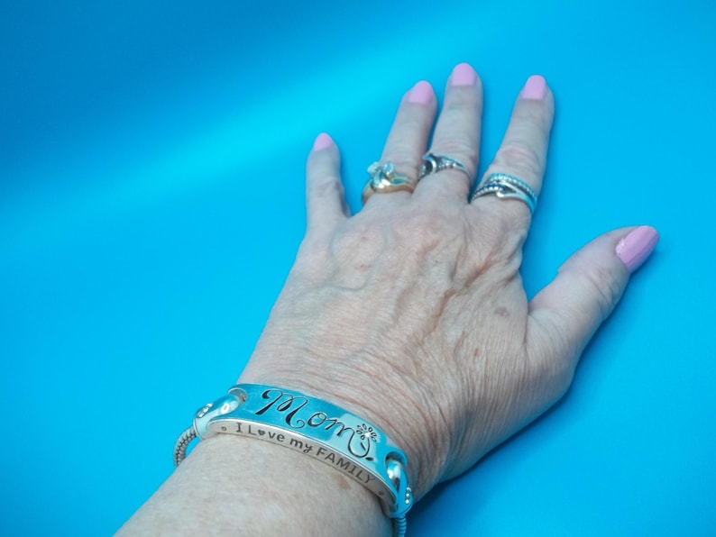 May include: A silver bracelet engraved with the word "Mom" and the phrase "I love my FAMILY". The bracelet is on a wrist with rings on the fingers. The background is a solid blue.