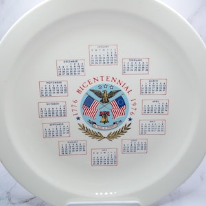 May include: White ceramic plate with a calendar design commemorating the Bicentennial of 1776. The plate features the text "Bicentennial 1776-1976" with an eagle, flags, and a bell. Calendars for each month are arranged around the central design.