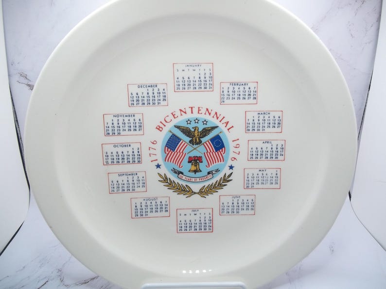 May include: White ceramic plate with a calendar design commemorating the Bicentennial of 1776. The plate features the text "Bicentennial 1776-1976" with an eagle, flags, and a bell. Calendars for each month are arranged around the central design.