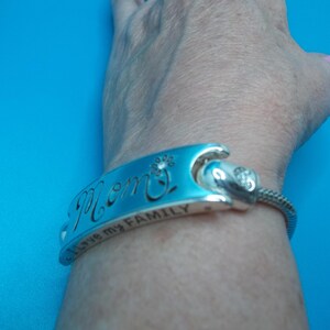 May include: A silver bracelet with the word "Mom" engraved on a blue enamel plate. The bracelet also has the inscription "I love my FAMILY". A small flower and a crystal accent the design.