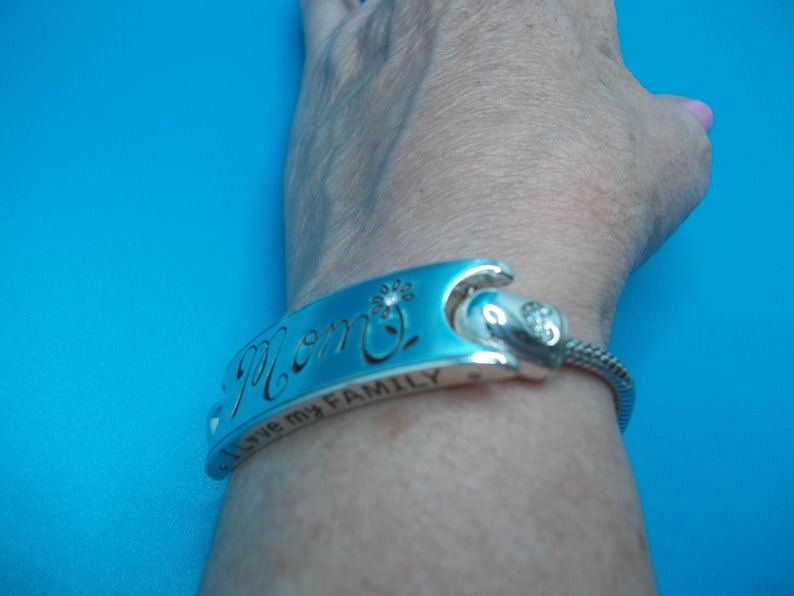 May include: A silver bracelet with the word "Mom" engraved on a blue enamel plate. The bracelet also has the inscription "I love my FAMILY". A small flower and a crystal accent the design.