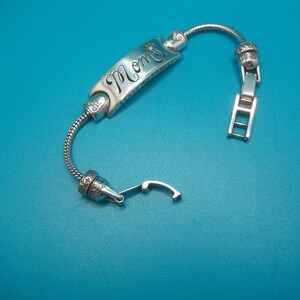 May include: Silver bracelet featuring the word "Mom" in cursive on a rectangular plaque. The bracelet has a flexible, snake-chain design and a clasp. The background is a teal colour.