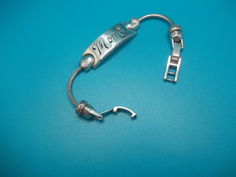May include: Silver bracelet featuring the word "Mom" in cursive on a rectangular plaque. The bracelet has a flexible, snake-chain design and a clasp. The background is a teal colour.