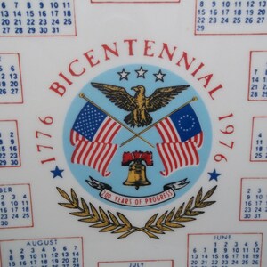 May include: A white ceramic plate with a calendar and a central emblem commemorating the Bicentennial of 1776. The emblem features an eagle, flags, and the Liberty Bell. The plate includes the years 1776 and 1976.
