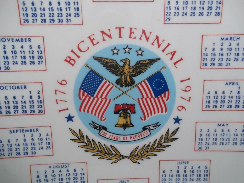 May include: A white ceramic plate with a calendar and a central emblem commemorating the Bicentennial of 1776. The emblem features an eagle, flags, and the Liberty Bell. The plate includes the years 1776 and 1976.