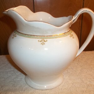 May include: White ceramic pitcher with a scalloped rim and a gold and green decorative band. The pitcher has a handle and a round base.
