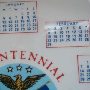 May include: A white ceramic plate with a calendar design featuring the months of January, February, and March. The plate has a red border around the calendar boxes and the word "CENTENNIAL" in red.