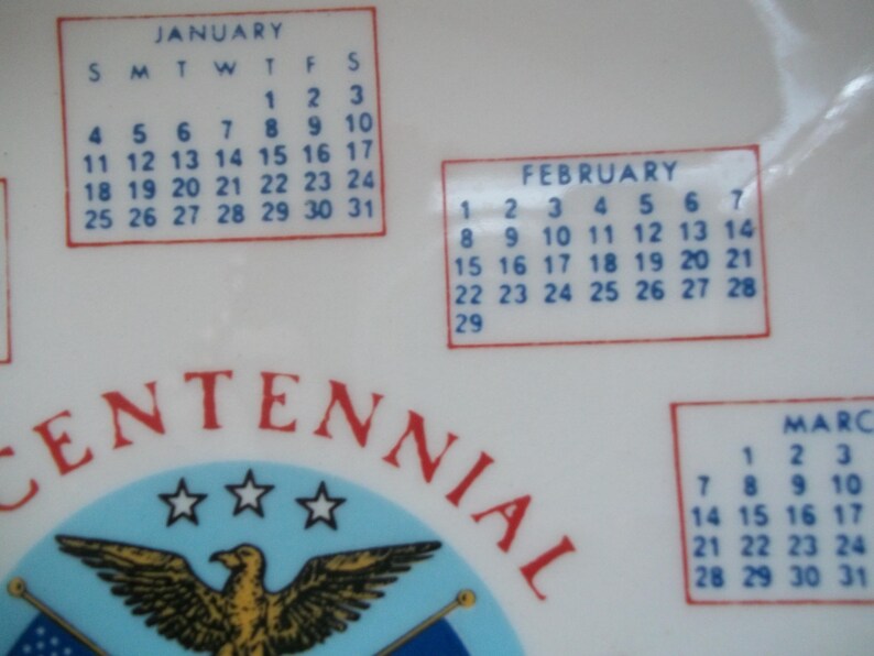 May include: A white ceramic plate with a calendar design featuring the months of January, February, and March. The plate has a red border around the calendar boxes and the word "CENTENNIAL" in red.