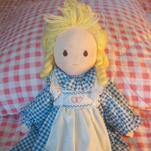 May include: A handmade doll with blonde hair wearing a blue and white gingham dress with a white apron and two red hearts embroidered on the front. The doll is lying on a pink and white checkered fabric.