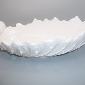 May include: A white, ceramic serving bowl with a leaf-like design. The bowl has a long, oval shape with a textured, scalloped edge and a rolled handle on one end. The bowl is approximately 14 inches long.