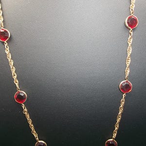 May include: A gold-toned necklace featuring a series of large, round, faceted red gemstones. The gemstones are evenly spaced along a delicate chain. The necklace is displayed against a dark background.