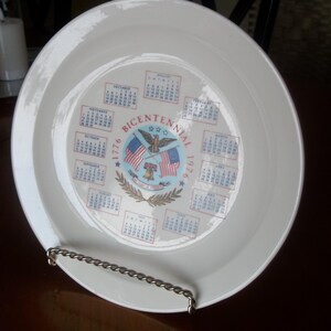 May include: A white ceramic plate featuring a Bicentennial design with the years 1776 and 1976. The plate's center displays an eagle, flags, and a calendar. The plate is held by a silver metal stand.