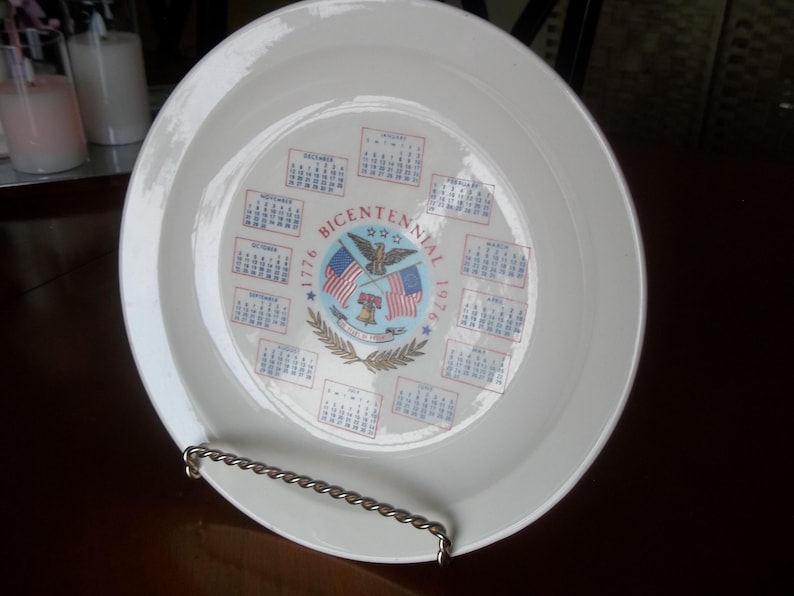 May include: A white ceramic plate featuring a Bicentennial design with the years 1776 and 1976. The plate's center displays an eagle, flags, and a calendar. The plate is held by a silver metal stand.