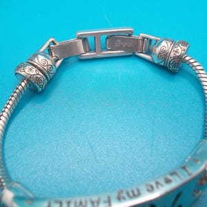 May include: Silver bracelet with floral and swirl detailing. The bracelet has a flexible, snake-like chain and a clasp with the brand name "Brighton". The bracelet has the words "I love my family" engraved on it.