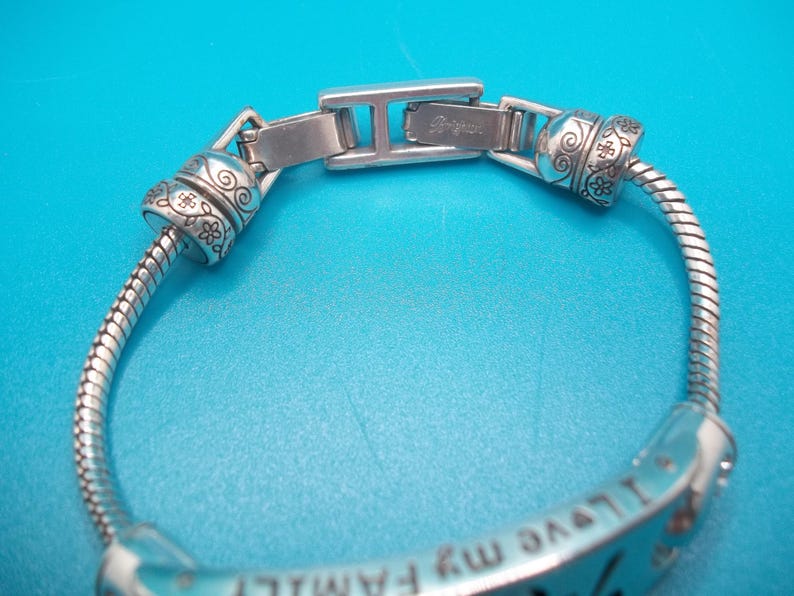 May include: Silver bracelet with floral and swirl detailing. The bracelet has a flexible, snake-like chain and a clasp with the brand name "Brighton". The bracelet has the words "I love my family" engraved on it.