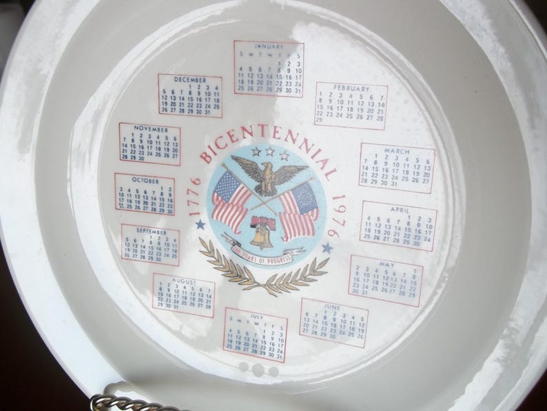 May include: A white ceramic plate featuring a calendar design for the year 1976, commemorating the Bicentennial. The design includes the American flag, an eagle, and the words "Bicentennial 1776-1976".