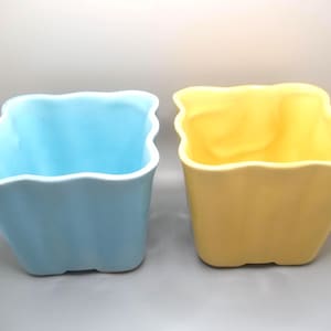May include: Two ceramic square planters, one light blue and one yellow, with a wavy rim design. The planters are empty and sit side-by-side on a gray surface. These decorative pots are suitable for indoor or outdoor use.