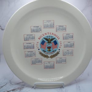 May include: Cream-colored commemorative plate with a calendar design. The center features a patriotic emblem with flags, an eagle, and the text "Bicentennial 1776-1976." Calendars for each month are arranged around the edge of the plate.
