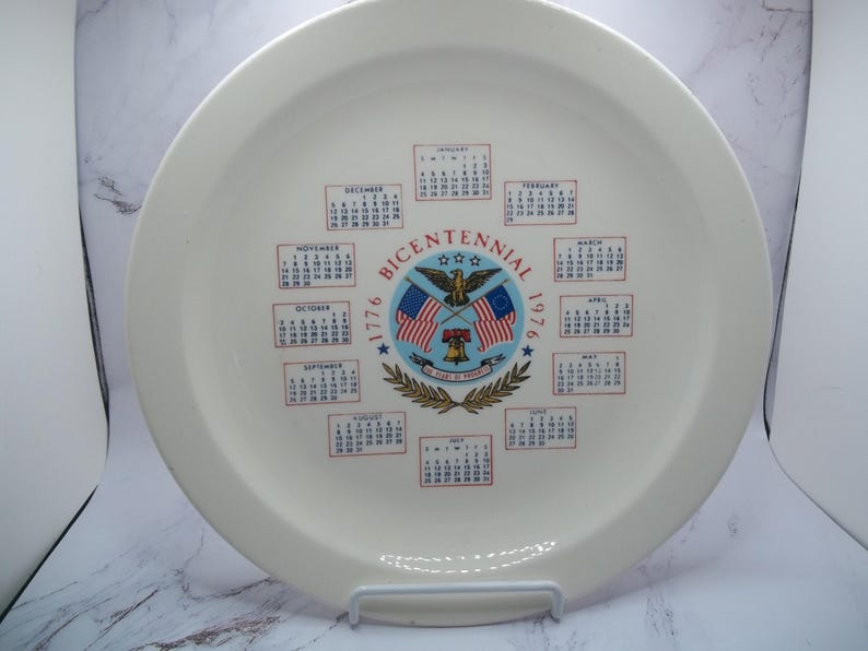 May include: Cream-colored commemorative plate with a calendar design. The center features a patriotic emblem with flags, an eagle, and the text "Bicentennial 1776-1976." Calendars for each month are arranged around the edge of the plate.